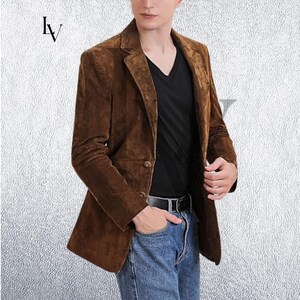 May include: A brown suede blazer with a notched lapel and two-button closure. The jacket is worn over a black V-neck shirt and blue jeans. The blazer has a classic, tailored fit.