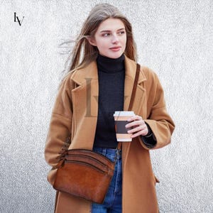 May include: A brown leather crossbody bag with three zippered compartments. The bag is worn over the shoulder. The person is wearing a brown coat, a black turtleneck, and holding a coffee cup. The background is a textured, light gray wall.