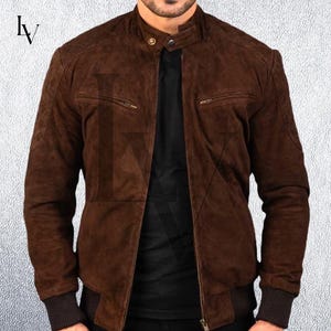 May include: A brown suede bomber jacket with a stand-up collar and two zippered pockets. The jacket has ribbed cuffs and a ribbed hem. The jacket is a medium brown color and has a soft, textured appearance.