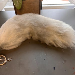 May include: A fluffy, light gray faux fur tail with a gold-colored keychain and clasp attached. The tail is long and tapered, with a soft, textured appearance. The keychain is round and has a small clasp for easy attachment.