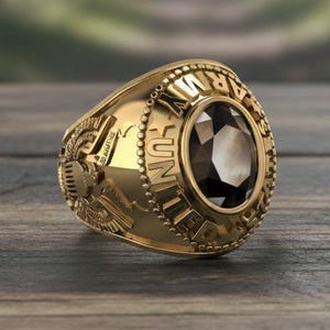 May include: A gold-colored ring with a dark, faceted center stone. The ring features the words "UNITED STATES ARMY" around the stone and detailed side emblems. The ring is set on a wooden surface.