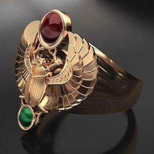 May include: Gold-colored ring featuring an Egyptian scarab beetle design. The ring has a red gemstone at the top and a green gemstone at the bottom. The beetle is centered on a winged design.