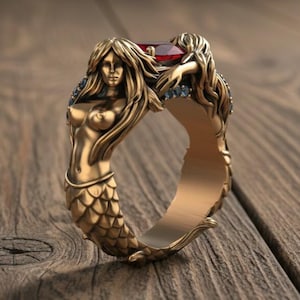 May include: A gold-colored ring featuring two mermaid figures. The mermaids have flowing hair and are positioned on either side of a red gemstone. The ring is set against a wooden background.