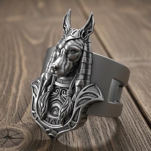 Może przedstawiać: Srebrna bransoletka mankietowa z szczegółowym wizerunkiem Anubisa, egipskiego boga z głową psa. Bransoletka jest umieszczona na drewnianym tle. Postać Anubisa jest misternie szczegółowa.