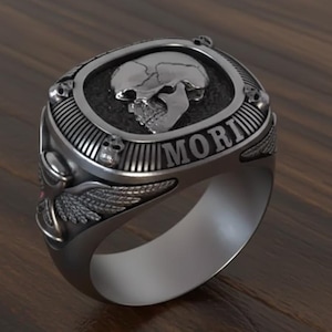 May include: A silver-toned ring with a skull design. The ring features a central oval with a skull, the word "MORI", and skull accents. Wings are on the sides of the ring.