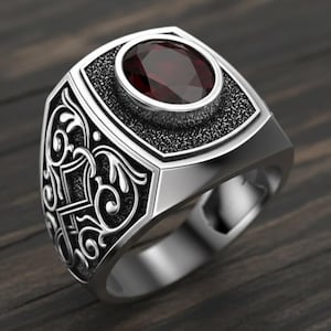 Op de afbeelding: Een zilveren ring met een grote, ovale, donkerrode edelsteen. De ring heeft een gedetailleerd, getextureerd ontwerp rond de edelsteen en aan de zijkanten. De ring is geplaatst tegen een donkere houtnerf achtergrond.