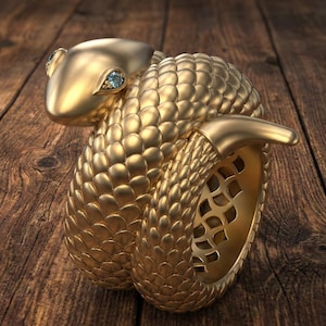May include: A gold snake ring with a detailed scale design. The snake's head has two small, light blue gemstones. The ring is displayed on a dark wood grain surface, showcasing the intricate details of the jewellery.