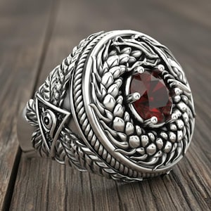 May include: A silver ring with intricate detailing and a large, round, red gemstone at the center. The ring features a detailed design around the gemstone and on the band, including a triangle with an eye symbol.