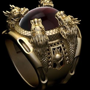 May include: A gold-colored ring featuring three dragon figures encircling a large, round, dark red gemstone. The ring has intricate details, including dragon scales and decorative patterns, set against a black background.