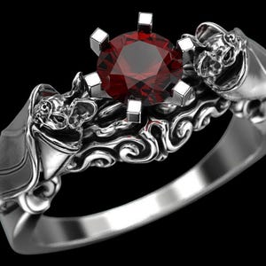 May include: A silver ring with a dark red gemstone at the center. The ring features bat-shaped designs on each side of the gemstone and intricate detailing along the band. The ring is set against a black background.