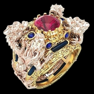 Puede incluir: Anillo dorado ornamentado con una gran gema roja facetada en el centro, rodeada de gemas azules más pequeñas. El anillo tiene detalles intrincados, incluyendo cabezas de dragón esculpidas y una banda decorativa. Fondo negro.