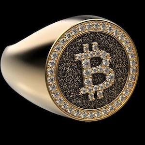 Puede incluir: Un anillo dorado con un símbolo de Bitcoin incrustado con pequeñas piedras brillantes. El símbolo de Bitcoin está centrado en un fondo texturizado marrón oscuro, rodeado por un anillo de piedras más pequeñas. El anillo tiene una banda lisa y pulida.