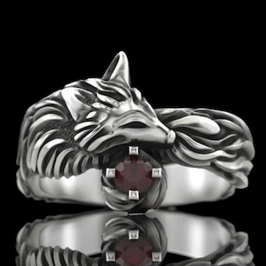 May include: A silver-colored ring featuring a detailed wolf design. The wolf's head is at the top, with a red gemstone set in the center. The ring has a polished finish and is set against a black background.