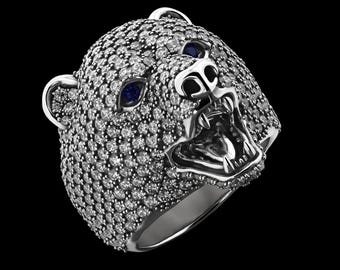 May include: A silver ring featuring a bear's head design. The bear's head is covered in small, sparkling stones, with blue stones for the eyes. The bear's mouth is open, revealing teeth. The ring is set against a black background.