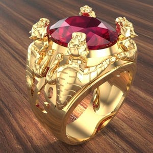 Puede incluir: Un anillo de oro con una gran gema roja facetada. La banda del anillo está adornada con tres figuras esculpidas. El anillo está sobre una superficie de madera, resaltando su intrincado diseño y artesanía.
