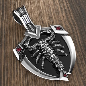 Könnte beinhalten: Silberner Anhänger mit detailliertem Skorpion-Design auf schwarzem Hintergrund. Der Anhänger hat eine schildförmige Form mit roten Edelsteinakzenten und einer dekorativen Öse. Der Skorpion ist das zentrale Element dieses Schmuckstücks.
