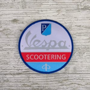 Puede incluir: Un parche redondo con el logotipo de Vespa en plata y la palabra "SCOOTERING" en rojo. El parche tiene un borde azul y un fondo blanco con un emblema azul y plateado.