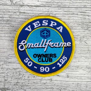 May include: A round, embroidered patch with a blue and yellow design. The patch features the word "VESPA" at the top, "Smallframe" in the center, and "OWNERS CLUB" below. Numbers 50, 90, and 125 are also included.