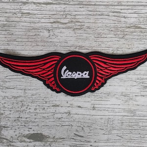 May include: A black and red embroidered patch featuring the Vespa logo within a circle, flanked by red wings. The Vespa text is in white. The patch is designed to be sewn or ironed onto fabric.