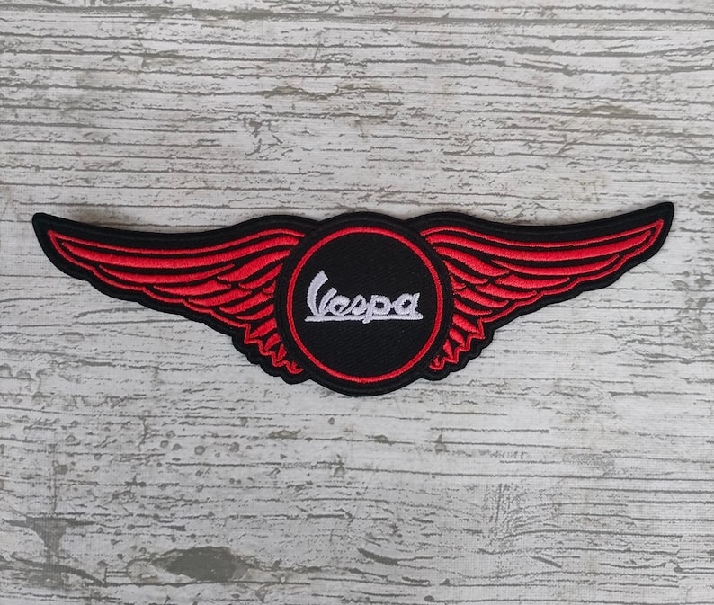 May include: A black and red embroidered patch featuring the Vespa logo within a circle, flanked by red wings. The Vespa text is in white. The patch is designed to be sewn or ironed onto fabric.