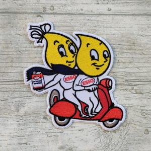 May include: Embroidered patch featuring two yellow cartoon characters riding a red scooter. One character holds a small container. Both characters are wearing white outfits with the word "ESSO" on them. The patch has a white border.