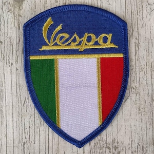 May include: Embroidered patch featuring the Vespa logo in gold script above the Italian flag. The patch has a blue shield shape with a gold border and a stitched edge. The flag colors are green, white, and red.