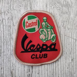 Op de afbeelding: Rode Vespa Club-patch met geborduurde details. Voorzien van het Castrol-logo, een scooterrijder en het woord "Vespa" in zwart schrift. De patch heeft een beige rand.
