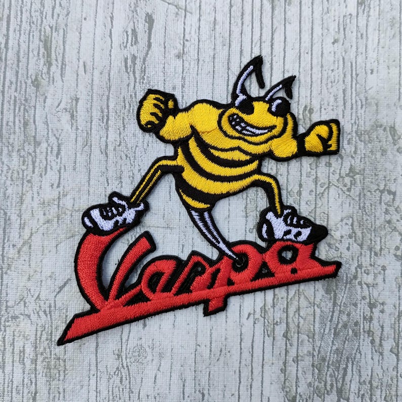 May include: Embroidered patch featuring a yellow and black cartoon bee with a smiling face, wearing sneakers, and the red word "Vespa". The bee is in a running pose, with its arms raised. The patch is on a light gray wooden surface.