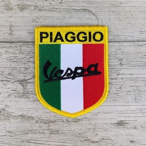 Puede incluir: Parche bordado con el logotipo de Vespa y la palabra "PIAGGIO" en negro sobre fondo amarillo. El parche tiene forma de escudo con los colores de la bandera italiana: verde, blanco y rojo.