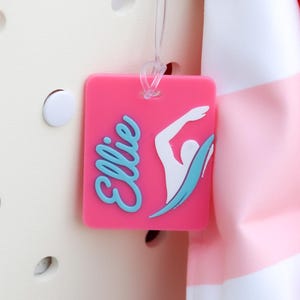 May include: A cream-colored perforated tote bag with a pink rectangular luggage tag attached. The tag features the name Ellie in blue script and a white graphic of a swimmer. A pink and white striped towel hangs over the side of the bag.
