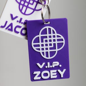 May include: A purple and white VIP Zoey keychain with a geometric design. The keychain has a rectangular tag with the text "V.I.P. ZOEY" in white. A second tag with the text "V.I. JACO" is also visible. The keychain is attached to a silver metal ring.
