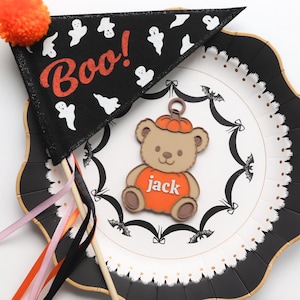 May include: A Halloween-themed decorative plate with a black and white scalloped edge. A black pennant with the word "Boo!" and ghost graphics is on top. A bear-shaped charm with a pumpkin hat and the name "jack" is also on the plate.