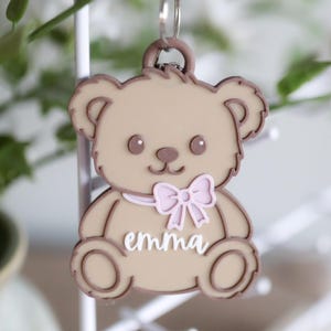 May include: A tan teddy bear keychain with a pink bow and the name "emma" in white script. The bear has brown outlines and details, including the ears, eyes, nose, and paws. The keychain is attached to a silver ring.