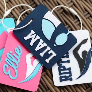 May include: Four rectangular luggage tags with white attachment loops. One is light blue with the name AVERY, one is pink with the name Ellie and a swimmer graphic, one is dark blue with the name LIAM and goggles, and one is white with RIPTIDE.