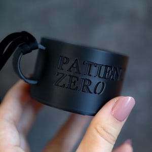 May include: A matte black cylindrical tag or band featuring the embossed text PATIENT ZERO in a bold, sans-serif font. The object has a smooth, solid finish and includes a small black loop attachment on the side for hanging or securing to items.