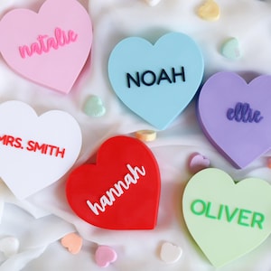Puede incluir: Una colección de decoraciones en forma de corazón en varios colores pastel, cada una con un nombre. Los corazones son rosas, azules, morados, blancos, rojos y verdes. Los nombres están escritos en un color contrastante. Caramelos decorativos y bolas de fieltro rodean los corazones.