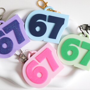 May include: A selection of keyrings in various colours, each displaying the number '67'. The keyrings are in shades of purple, blue, pink, and green, with silver and gold-coloured clasps. The numbers are in contrasting colours.