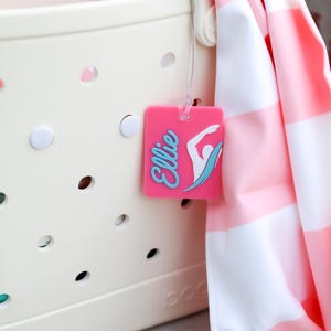 May include: A cream-colored perforated tote bag with a pink rectangular luggage tag attached. The tag features the name Ellie in blue script and a white graphic of a swimmer. A pink and white striped towel hangs over the side of the bag.