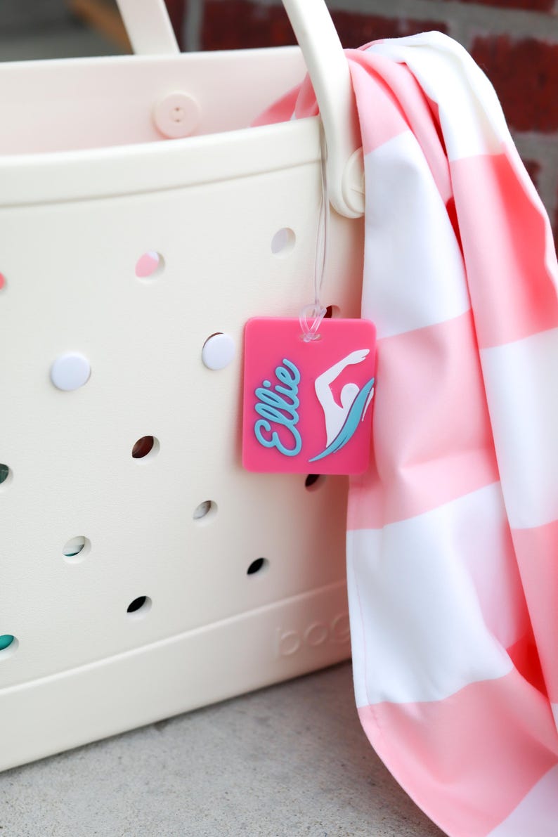 May include: A cream-colored perforated tote bag with a pink rectangular luggage tag attached. The tag features the name Ellie in blue script and a white graphic of a swimmer. A pink and white striped towel hangs over the side of the bag.