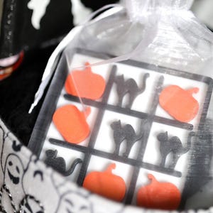 May include: A Halloween-themed Tic-Tac-Toe game with orange pumpkin and black cat game pieces. The game is in a clear bag tied with a white ribbon, inside a Halloween-themed bag with ghost and jack-o'-lantern designs.