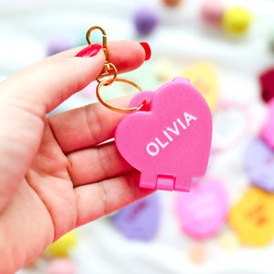 May include: A pink heart-shaped keychain with the name "OLIVIA" in white lettering. The keychain is attached to a gold-colored clasp and ring. The background features other heart-shaped items and colorful pom-poms.