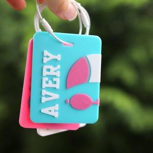 May include: A rectangular luggage tag in bright turquoise featuring the name AVERY in raised white letters. To the right of the name is a pink and white graphic of a hot air balloon. The tag is attached to a white wire loop for secure fastening.