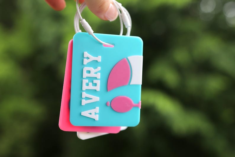 May include: A rectangular luggage tag in bright turquoise featuring the name AVERY in raised white letters. To the right of the name is a pink and white graphic of a hot air balloon. The tag is attached to a white wire loop for secure fastening.