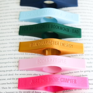 May include: A collection of colorful, plastic book page holders in various colors, each with a different engraved message. The messages include "FOR OUR NEXT CHAPTER", "FOR MY FAVORITE BOOKWORM - DAD", and "MAIN CHARACTER ENERGY".
