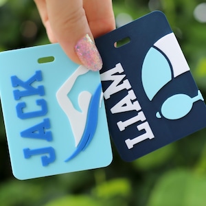 May include: Two rectangular luggage tags with rounded corners. One light blue tag features the name JACK in blue letters and a white swimmer graphic. The other dark blue tag features the name LIAM in white letters and a white and light blue graphic.