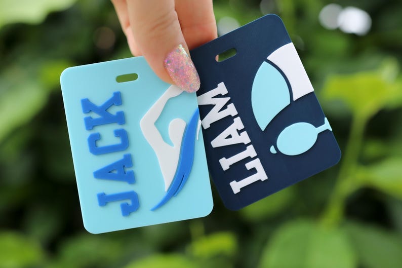 May include: Two rectangular luggage tags with rounded corners. One light blue tag features the name JACK in blue letters and a white swimmer graphic. The other dark blue tag features the name LIAM in white letters and a white and light blue graphic.