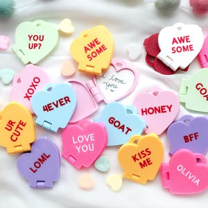 May include: A collection of colorful heart-shaped lockets with various phrases like "YOU UP?", "4EVER", and "KISS ME". The lockets are in shades of pink, yellow, blue, and purple, scattered on a white surface with decorative pom-poms.