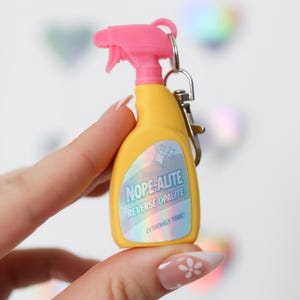 May include: A yellow spray bottle keychain with a pink trigger. The bottle has the text "NOPE-ALITE REVERSE OPALITE EXTREMELY TOXIC" in blue and white. The keychain is attached to a silver ring.
