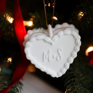 May include: A white heart-shaped Christmas ornament with a scalloped edge and the initials "M & S" in the center. The ornament hangs from a gold string against a backdrop of a Christmas tree with red ribbon and twinkling lights.