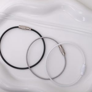 May include: Three circular cable key rings in black, silver, and white. Each ring features a thin, flexible metal wire construction with a threaded, cylindrical screw-lock closure mechanism made of metal for secure fastening of keys or tags.